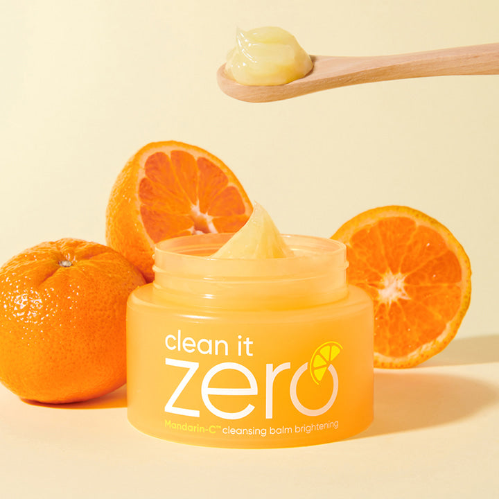 An open jar of BANILA CO - Clean It Zero Cleansing Balm Brightening 100ml, renowned as a waterproof makeup remover, sits on a surface surrounded by three mandarins. A wooden spoon with a small amount of the brightening balm is held above the jar. The label highlights "Mandarin-C cleansing balm.