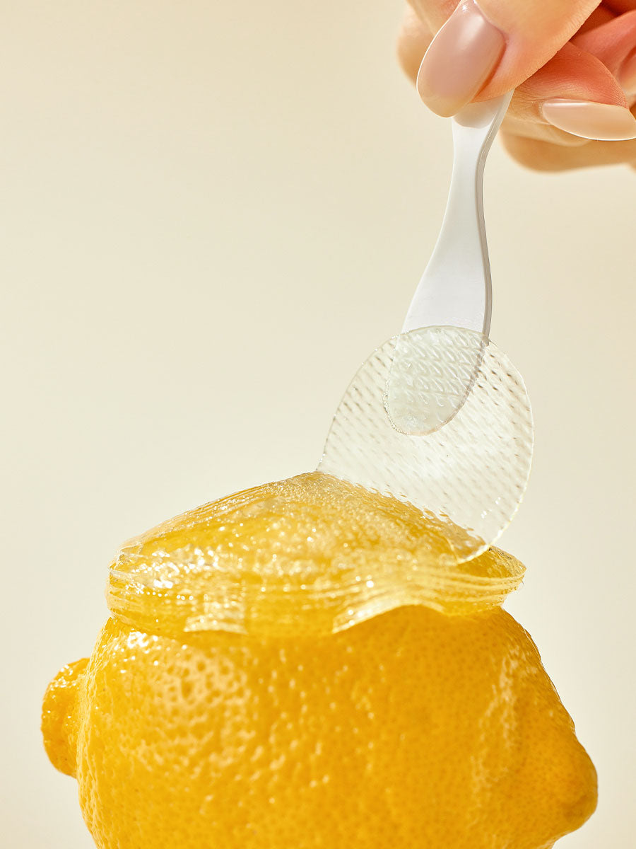 A hand holds a white plastic spoon, peeling back a transparent hydrogel sheet from the top of a fresh yellow lemon. The background is a plain, light beige color. The image focuses on the texture and color of the lemon and the clear layer, resembling GOODAL - Green Tangerine VitaC Eye Gel Patch 60ea by GOODAL.