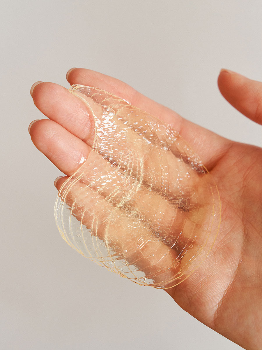 A hand is holding a transparent, jelly-like hydrogel sheet mask with a textured surface. The background is plain and neutral, allowing focus on the mask and hand. The fingers are gently pinching the mask, showcasing its flexible and delicate nature, perfect for 5-minute hydration under eye circles. GOODAL - Green Tangerine VitaC Eye Gel Patch 60ea by GOODAL.