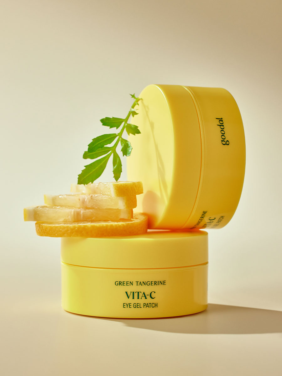 The image displays two yellow containers of "GOODAL - Green Tangerine VitaC Eye Gel Patch 60ea" by GOODAL, stacked atop each other. Fresh lemon slices and a green herb are placed next to the containers, emphasizing a fresh, citrusy theme with 5-minute hydration to combat under eye circles.