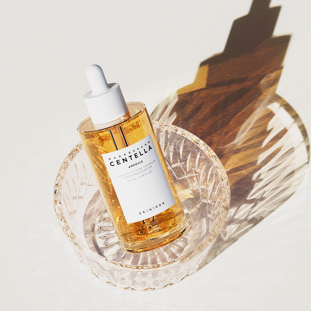 A clear glass dropper bottle of SKIN1004 - Madagascar Centella Ampoule 100ml, brimming with soothing ampoule, rests in a decorative crystal dish on a white surface. The sunlight casts shadows, highlighting the texture of the golden liquid and the intricate dish, emphasizing its potential to strengthen your skin barrier.
