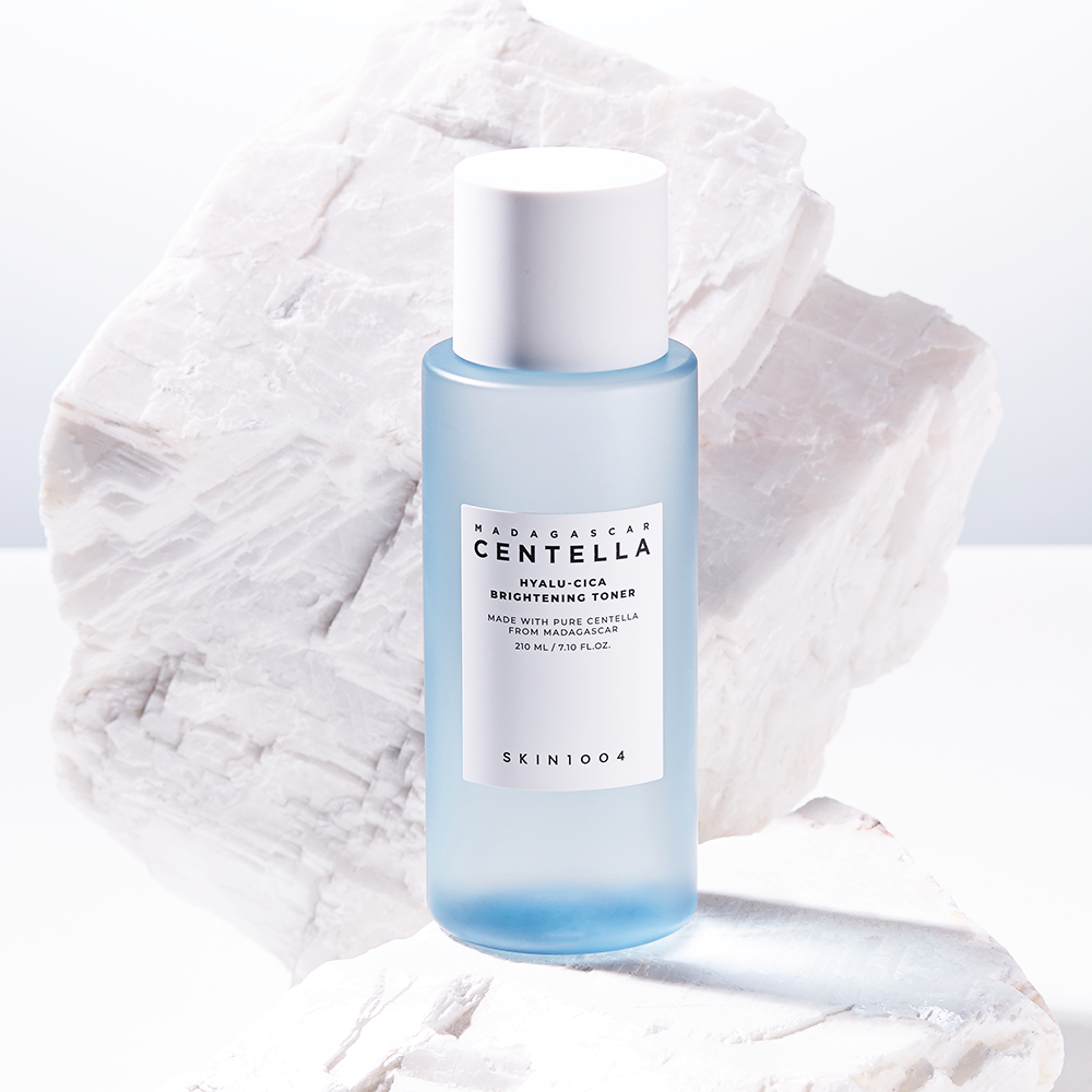 A frosted blue bottle of SKIN1004 - Madagascar Centella Hyalu-Cica Brightening Toner 210ml, a super-hydrating toner with AHA, is positioned in front of white marble blocks. The bottle has a white label and white cap, containing 210 ml (7.1 fl. oz.) of toner made with Centella Asiatica from Madagascar.
