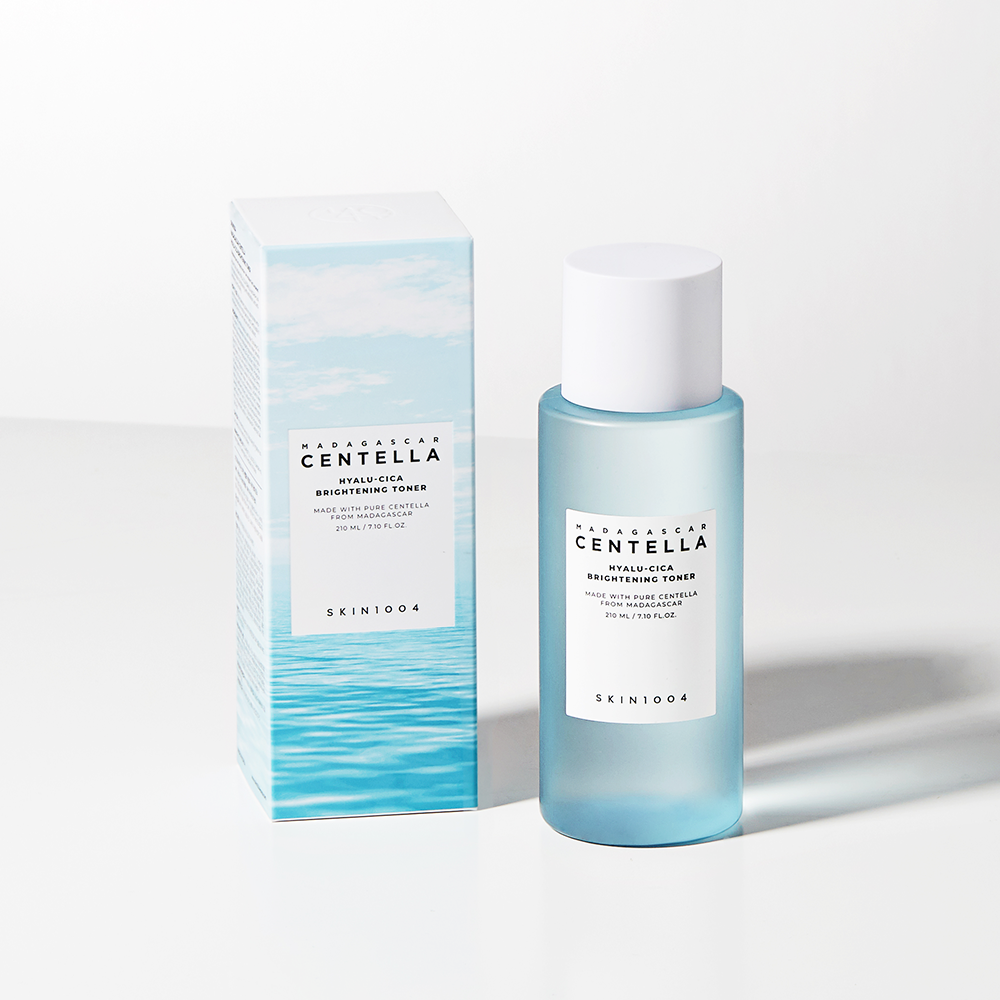 A bottle of SKIN1004 - Madagascar Centella Hyalu-Cica Brightening Toner 210ml stands next to its box. This super-hydrating toner features a frosted bottle with a white cap and light blue gradient design, while the box exhibits a matching blue gradient. Both items rest elegantly on a white surface.