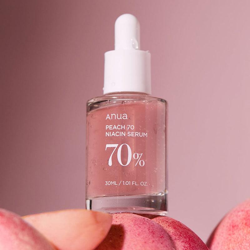 A glass dropper bottle of ANUA - Peach 70% Niacinamide Serum 30ml is displayed against a soft pink background. The bottle is filled with a pinkish liquid and labeled with "70%" indicating its concentration. At the base of the image, blurred peach fruits hint at the peach extract. The bottle is 30ml or 1.01 fl oz.