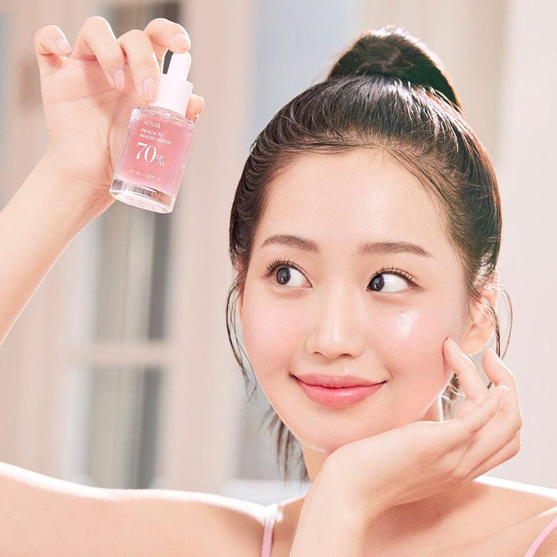 A woman with her hair in a bun is smiling and holding up a small bottle of ANUA - Peach 70% Niacinamide Serum 30ml in her left hand. The bottle, labeled "70%", is filled with a pink liquid, likely infused with peach extract. She gazes at it as sunlight filters through a window in the background, giving her glass skin an ethereal glow.