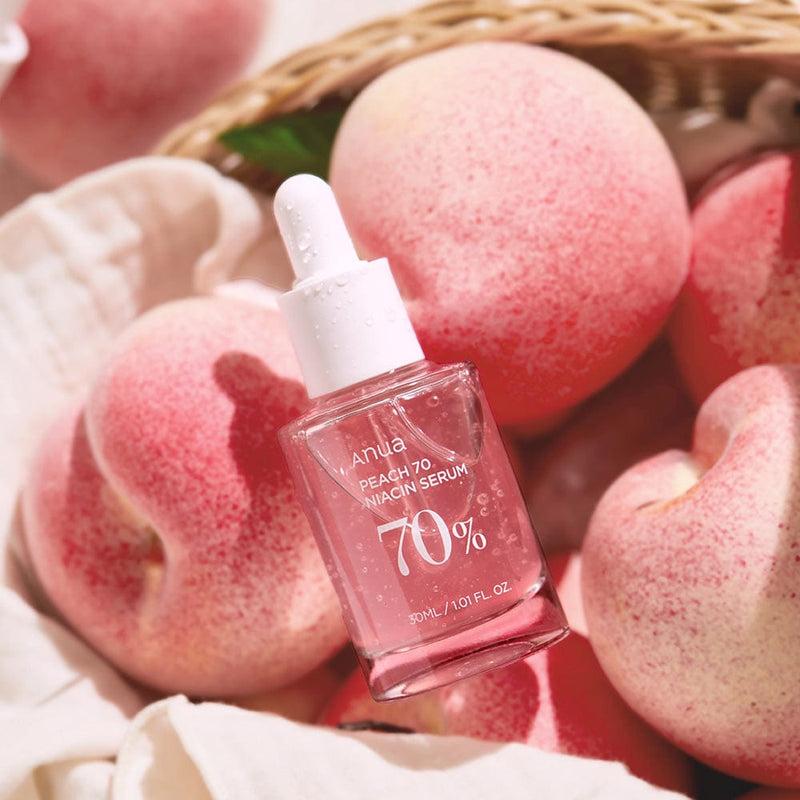A bottle of ANUA - Peach 70% Niacinamide Serum 30ml is placed among fresh peaches. The clear glass bottle has a white dropper lid and contains pink liquid. The label shows the product name and '70%' clearly, highlighting its rich peach extract. The background includes a wicker basket and peach-colored fabric, perfect for achieving that coveted glass skin look.