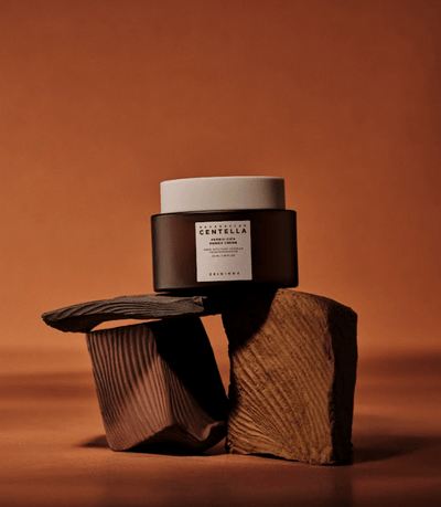 A small jar of SKIN1004 - Probio-Cica Enrich Cream 50ml with a white lid is placed on two textured, brown wooden blocks against a brown background. The design gives a minimalistic and natural aesthetic, perfectly suited for those seeking to strengthen their skin barrier.