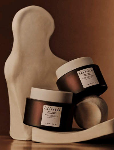 Two brown skincare jars with white labels and white lids are balanced on a sculptural, abstract white object against a warm, brown background. The labels read "Probio-Cica Enrich Cream 50ml" by SKIN1004. The artistic and minimalist arrangement highlights this moisturizing cream for the skin barrier.