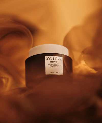 A brown jar with a white cap containing moisturizing cream is centered in the image. The label reads "SKIN1004 - Probio-Cica Enrich Cream 50ml" and includes other text. The background is an abstract mix of warm orange and brown tones, creating a glowing effect around the jar.