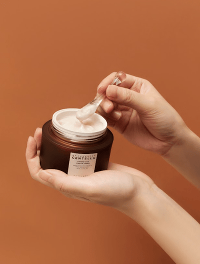 A person holding an open jar of SKIN1004 - Probio-Cica Enrich Cream 50ml with one hand while using a small clear spatula to scoop some product with the other hand. The background is a solid, warm orange color, highlighting the importance of maintaining your skin barrier.