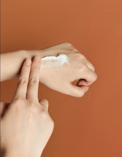 A person is applying a dab of SKIN1004 - Probio-Cica Enrich Cream 50ml, enriched with Centella Asiatica, onto the back of their hand using their index and middle fingers. The background is a solid orange color.