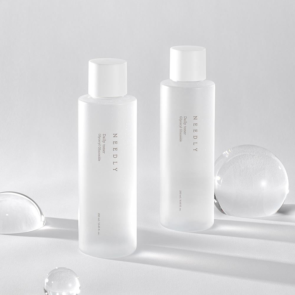 Two translucent white bottles of NEEDLY - Daily Toner 250ml stand on a white surface. The bottles are simple in design, with white caps and minimalist text. Several clear, smooth spheres are placed around the bottles, reflecting light and adding a stylish touch to the scene—perfectly complementing this mild exfoliating and moisturizing toner.