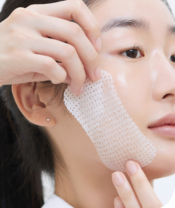 A person with fair skin and dark hair is applying a MEDIHEAL - Retinol Collagen Lifting Pad 100ea to their cheek. The close-up profile view highlights the process of addressing fine lines and wrinkles, with the pad held by their hand near the cheekbone.