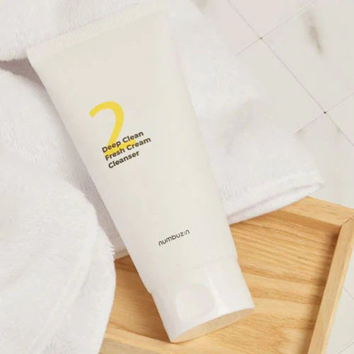 A white tube of "Numbuzin - No.2 Deep Clean Fresh Cream Cleanser 120ml" sits elegantly on a wooden tray. Designed for sensitive skin, this cleansing foam makes pore waste a thing of the past. A pristine white towel rests in the background, set against the minimalist tiled surface in the upper right corner.