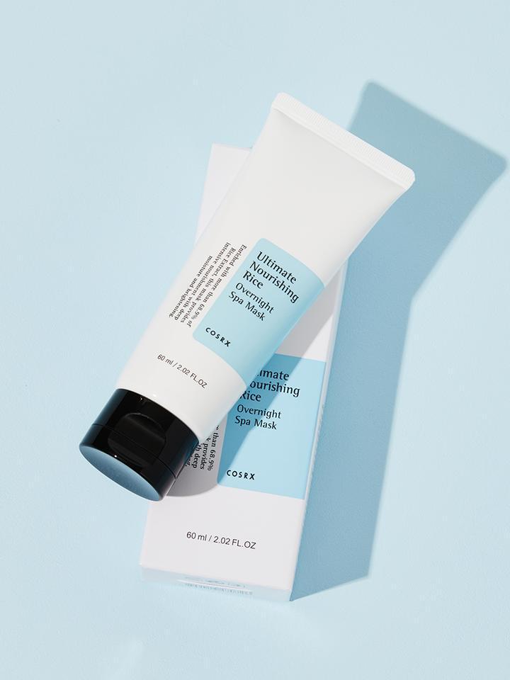 A white tube labeled "COSRX - Ultimate Nourishing Rice Overnight Spa Mask 60ml" is placed atop its matching blue and white box. The tube, featuring a black cap, is tilted to the right, casting a shadow on a light blue background. This 60 ml (2.02 fl. oz.) Rice Extract Mask promises nourishing and moisturizing benefits.