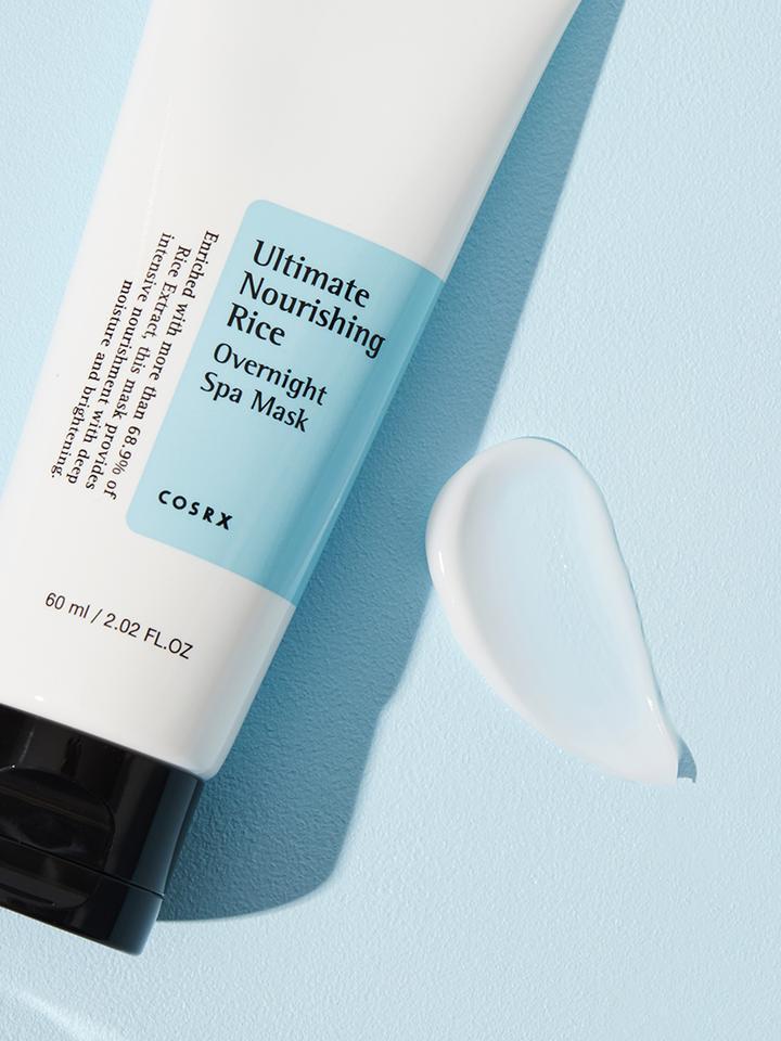 A white tube with a black cap labeled "COSRX - Ultimate Nourishing Rice Overnight Spa Mask 60ml" lays on a light blue surface. A small dollop of the nourishing and moisturizing rice extract mask is beside the tube. The product size is 60 ml (2.02 fl. oz.).