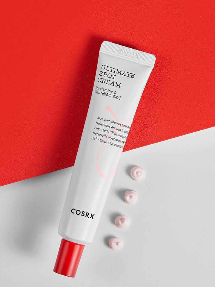 A tube of COSRX - AC Collection Ultimate Spot Cream 30g, an effective overnight acne solution, is placed diagonally on a two-tone red and white background. The white tube has red and black text and a red cap. Four small dollops of cream are placed in a line next to the tube.