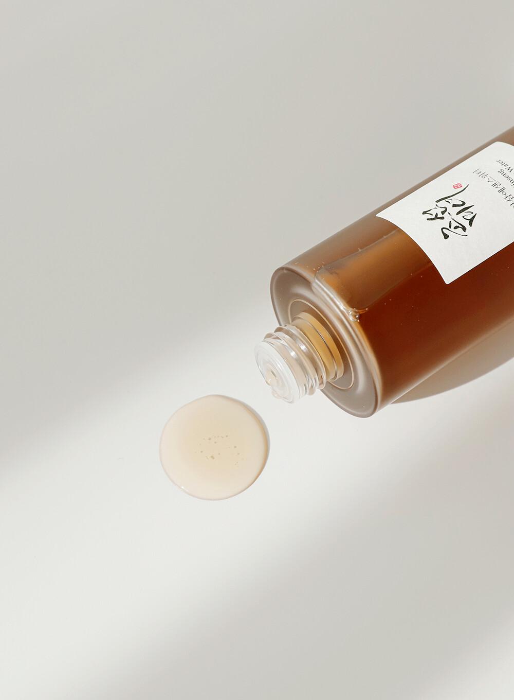 A bottle of BEAUTY OF JOSEON - Ginseng Essence Water 150ml is tilted, with a small amount of the brown liquid pooled at the opening. The bottle has a white label with some text and designs and is set against a clean, white background. This nutrient supply promises to offer hydrating skin benefits.