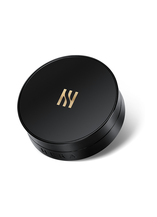 A sleek, round compact case with a black matte finish is shown. It features a gold "H" emblem on the lid and the brand name "HERA" subtly embossed along the side of the base. The case houses a luxurious liquid foundation perfect for achieving flawless coverage, specifically HERA - Black Cushion SPF 34 / PA++ - 15g (2ea).