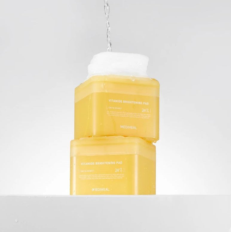 Two bright yellow containers of MEDIHEAL - Vitamide Brightening Pad 100ea, featuring the "VITAMIN C BRIGHTENING PAD" in white text, sit stacked on a white surface with water splashing onto the top. Infused with niacinamide, these glow-getter pads promise radiant skin and a minimalistic design.