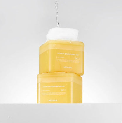 Two bright yellow containers of MEDIHEAL - Vitamide Brightening Pad 100ea, featuring the "VITAMIN C BRIGHTENING PAD" in white text, sit stacked on a white surface with water splashing onto the top. Infused with niacinamide, these glow-getter pads promise radiant skin and a minimalistic design.