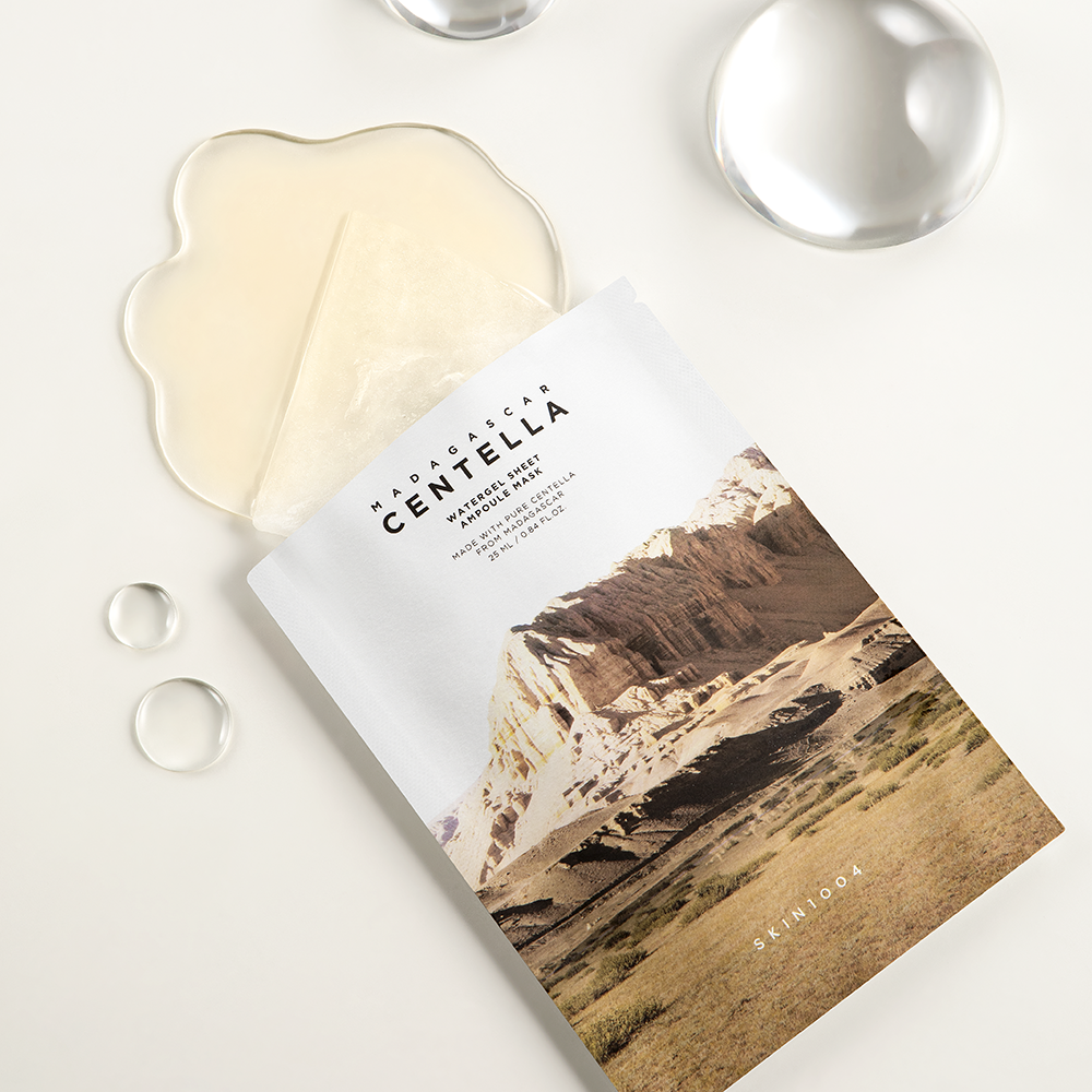 A flat lay photo of a skincare product showing a gel sheet mask partially pulled out from its packaging. The packaging features an image of a mountainous landscape and the text "SKIN1004 - Madagascar Centella Watergel Sheet Ampoule Mask 5ea." Infused with Centella Asiatica extract, it promises to intensely soothe and hydrate your skin. Clear droplets are scattered around the product.