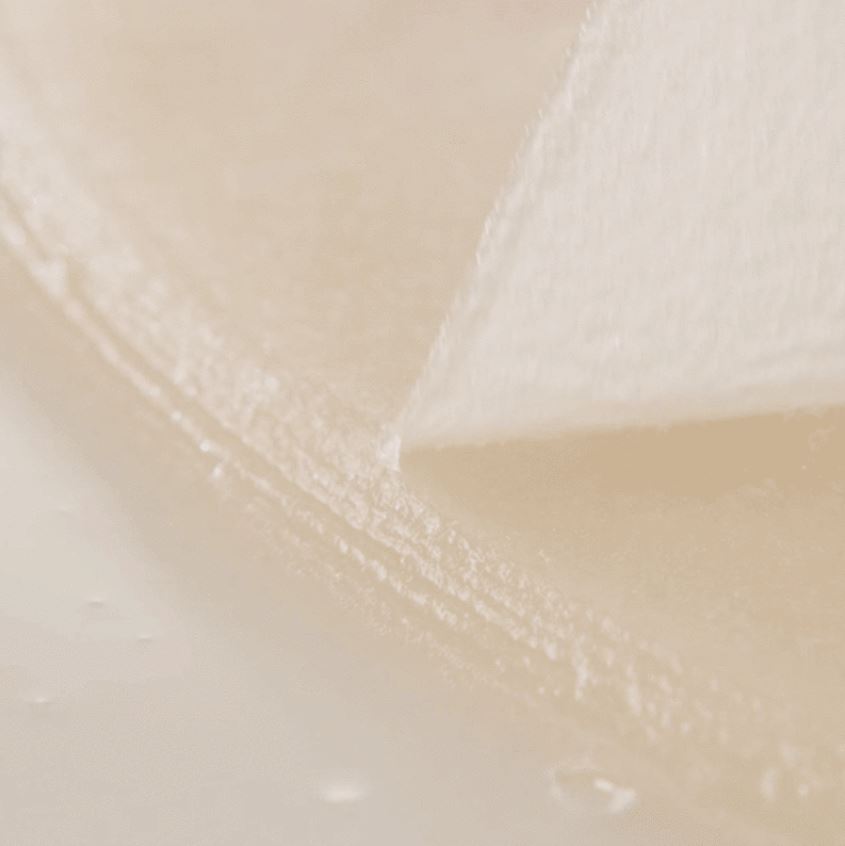 Close-up of a white, textured surface with a thin, angular object resting on it. The background is blurred, creating a soft, muted tone. The material, resembling an SKIN1004 - Madagascar Centella Watergel Sheet Ampoule Mask 5ea by SKIN1004, appears delicate with subtle lighting highlighting its texture.