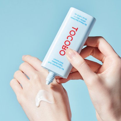 A person is applying TOCOBO - Bio Watery Sun Cream SPF50+ PA++++ 50ml from a blue and white tube labeled "Tocobo" onto the back of their hand. The background is a solid light blue. The cream, which boasts SPF 50+ protection, looks smooth and moisturizing.