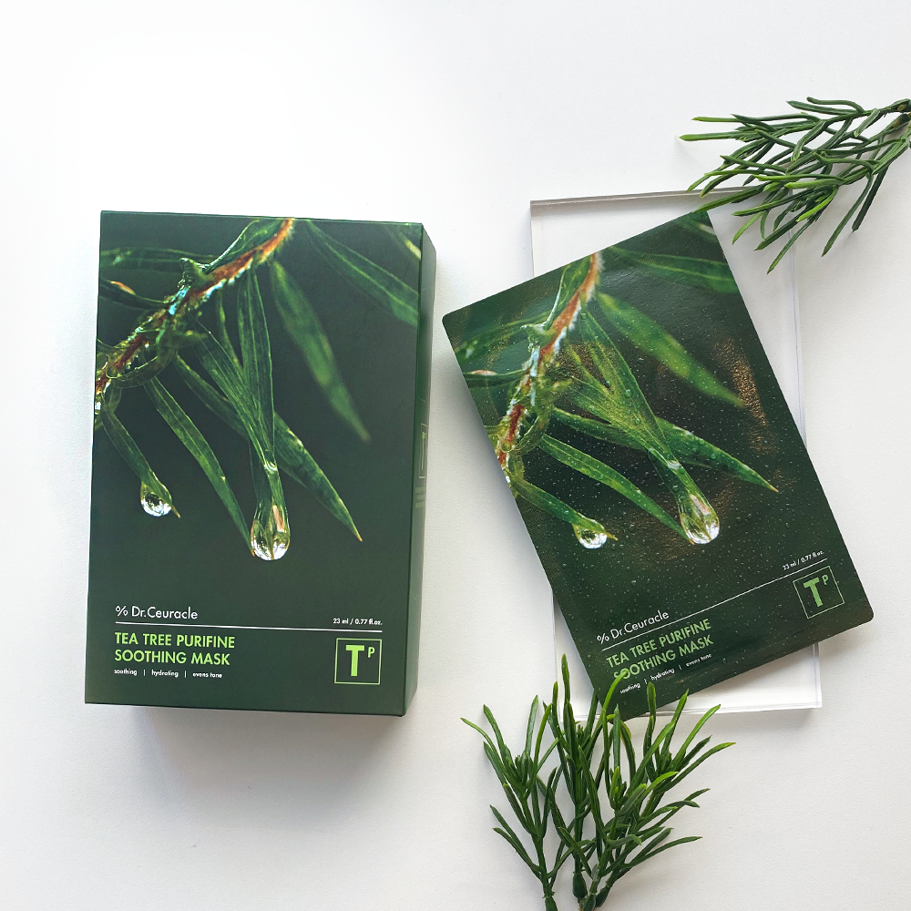 A box and an opened sachet of the "DR. CEURACLE - Tea Tree Purifine Soothing Mask 10pcs," infused with tea tree extract, are displayed on a white surface. The packaging showcases close-up images of tree leaves with droplets of water. Surrounding the products are sprigs of green foliage for decoration.