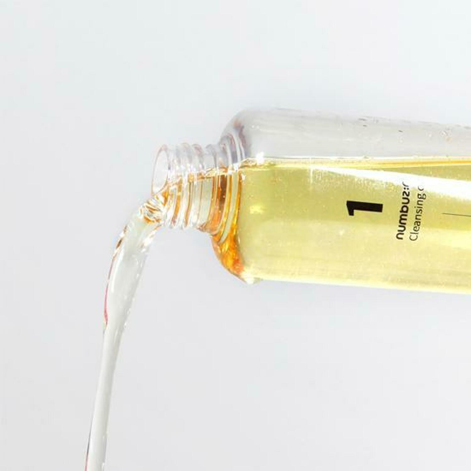 A close-up of a clear bottle labeled "1" with liquid flowing out of it. The light yellow substance appears to be a moisturizing cleanser formulated with naturally-derived oils. The cylindrical bottle is set against a plain white background, highlighting its simplicity and effectiveness. This product is the NUMBUZIN - No.1 Easy Peasy Cleansing Oil 200ml by Numbuzin.