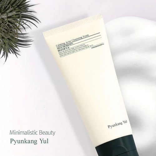 A white tube of PYUNGKANGYUL - Calming Acne Facial Cleanser 100ml, ideal for acne-prone skin, is placed against a light background. The tube features a dark green cap and minimalistic text. The brand name "PYUNKANGYUL" is visible. To the top left, there are some green plant fronds.