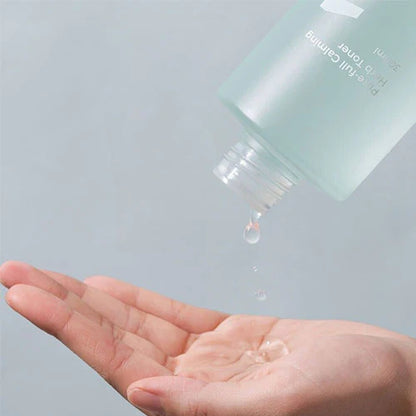 A close-up of a hand holding a few drops of liquid being dispensed from a frosted bottle with a blue hue and white text, containing NUMBUZIN - No.1 Pure-Full Calming Herb Toner 300ml by Numbuzin. The background is a plain, light, neutral color.