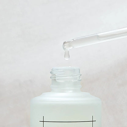 A close-up image of a dropper dispensing a clear liquid into the opening of a frosted glass bottle. The background is a soft, blurred white, highlighting the infusion that deeply hydrates and soothes sensitive skin with Centella Asiatica Extract from NUMBUZIN - No. 7 Mild Green Soothing Serum 50ml by Numbuzin.