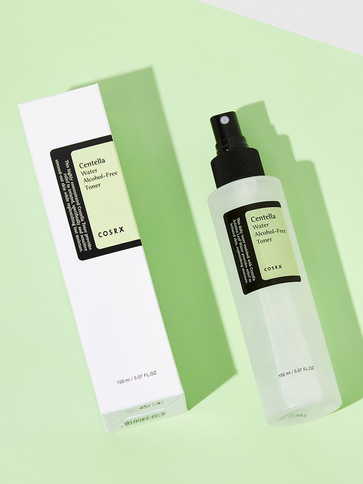 Image of a COSRX - Centella Water Alcohol-Free Toner 150ml. The toner, infused with Centella Asiatica from Jeju Island, is in a translucent bottle with a black spray cap, placed next to its white and light green box. The background is a light green surface, creating a fresh and clean ambiance that's both soothing & hydrating.