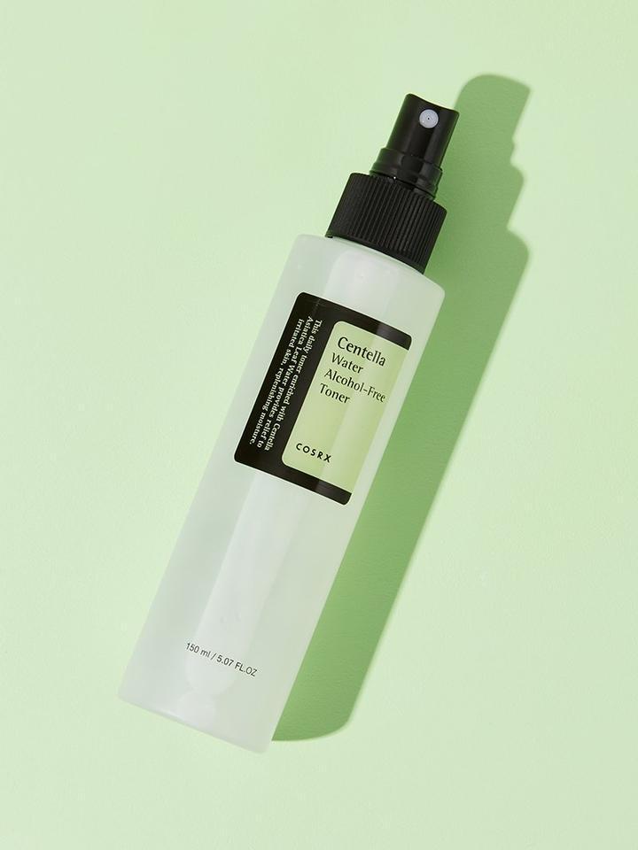 A clear spray bottle with a black label and spray nozzle, containing COSRX - Centella Water Alcohol-Free Toner 150ml. Enriched with soothing and hydrating Centella Asiatica from Jeju Island, the label features green and white text describing the product. The bottle is placed against a light green background, casting a shadow to the right.