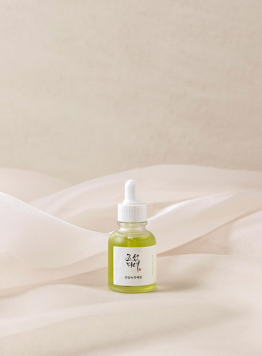 A small, transparent bottle with a white dropper cap containing a light-green liquid is set against a soft, beige fabric background. The label, in foreign characters, hints at ingredients like mugwort and green tea. This product is the BEAUTY OF JOSEON - Calming Serum Green tea + Panthenol 30ml from BEAUTY OF JOSEON.