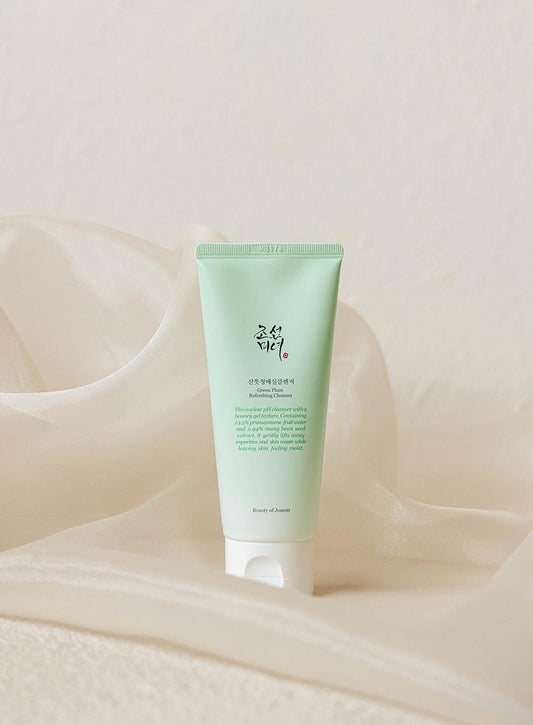 A light green tube of BEAUTY OF JOSEON - Green Plum Refreshing Cleanser 100ml with natural ingredients stands upright on a soft, white, silky fabric background. The tube has a white flip-top cap. The product name is visible but not legible in this image.