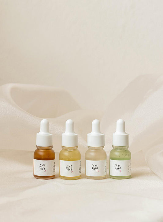 A mini size set of four small dropper bottles filled with different colored liquids, ranging from brown to green, is neatly lined up against a soft, light-colored fabric background. Each bottle from the BEAUTY OF JOSEON - Hanbang Serum Discovery Kit (10ml*4ea) has Korean text on its label and a white cap with a dropper.