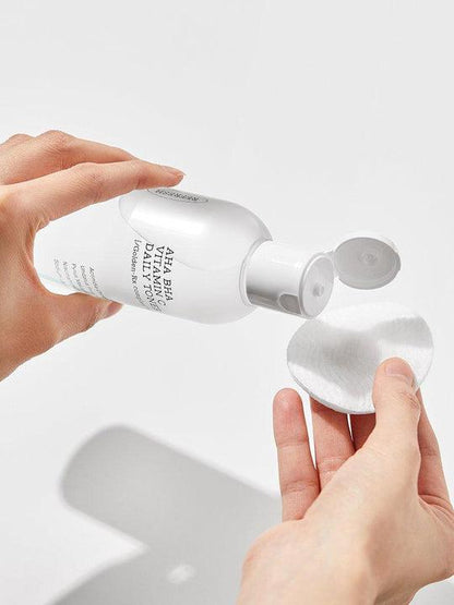 A person is holding a white plastic bottle labeled "COSRX - Refresh AHA BHA Vitamin C Daily Toner 280ml" and pouring its contents onto a round cotton pad, perfect for daily skin care. The bottle's cap is open, and the background is plain and white, highlighting the COSRX skincare product.