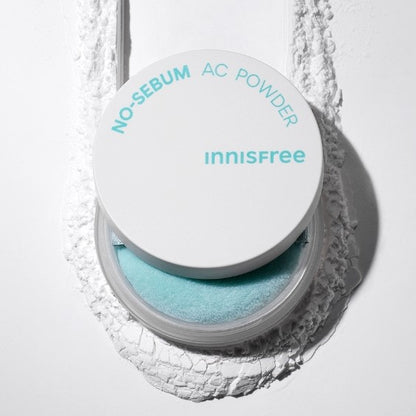 A container of INNISFREE - No-Sebum AC Powder 5g is shown. The lid of the round container is open, revealing a pale turquoise puff applicator. The background is textured with a white powdery substance, highlighting its hypoallergenic properties perfect for managing excessive sebum and troubled skin.