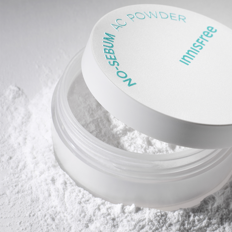 A container of INNISFREE - No-Sebum AC Powder 5g is open, revealing fine hypoallergenic powder inside. Some of the powder is scattered around the container on a white surface, ideal for absorbing excessive sebum and calming troubled skin. The product label is visible on the lid.