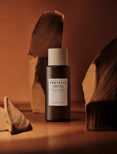 A skincare bottle labeled "SKIN1004 - Probio-Cica Toner 210ml" stands upright in the center, surrounded by abstract, earthy-toned rock-like formations. The backdrop is a warm, brown gradient, creating a natural and serene atmosphere. Infused with Centella Asiatica and ceramide NP, the bottle has a minimalistic white label and cap.