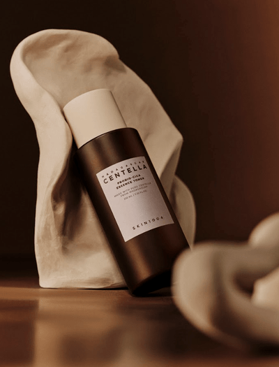 A brown bottle labeled "SKIN1004 - Probio-Cica Toner 210ml" with a white cap is positioned upright, supported by a sculptural, organic-shaped white form. Infused with Centella Asiatica and ceramide NP, the setting has warm lighting and a soft-focus effect, creating an elegant and minimalist aesthetic.