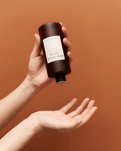 A person is tilting a brown bottle labeled "SKIN1004 - Probio-Cica Toner 210ml" over their open palm, allowing a liquid to dispense. The background is a solid, warm orange.